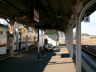 ホームを階段付近から縦に見て。吊られた駅名表、ホームに立つ時刻表、LED式発車案内板、待合室。