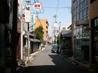 1.5車線の道路の脇に家屋や小さなビルが立ち並んでいる。アーケードはない。