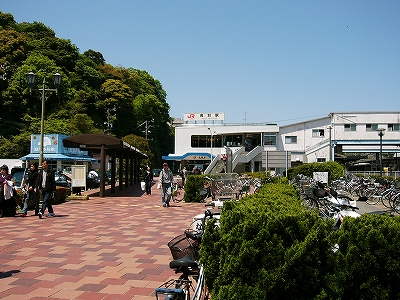 緑の植え込みに囲まれた屋根の無い駐輪所、そのかなり向こうに駅舎。