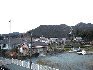 左手に家々が立ち並び、右手に砂利敷きの無機的なスペース。背景に低い里山の尾根。