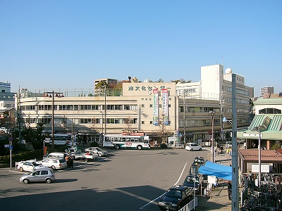 アスファルトの自由なスペースの右に橋上駅への階段、奥にはコーナーにあって横長に構えた古い建物。