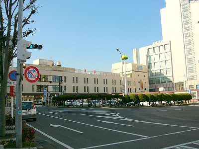 3階建ての横長の津駅駅舎。左手にアスト津の高い建物。駅舎前のロータリーは、背の低い緑のこんもりした木で囲われているようだ。