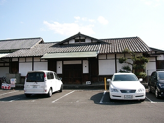 瓦屋根と白漆喰とも木柱の建物。こちらに流れてくる屋根には、こちらに向かって妻型の屋根が小さく突き出ている。