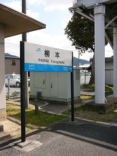 ホームに二本足で立つ駅名標。後ろにコンクリートの無機質な信号機室。