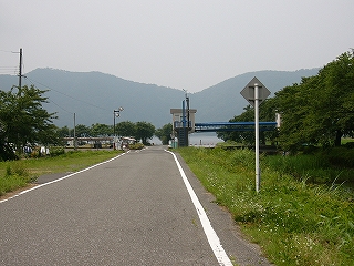 余呉湖に向かってまっすぐ伸びる道。車も人もない。右の道路わきには草が茂っている。ずっと奥の方に山々。