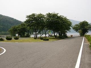 直進と左折に分かれた道別れ。股の間には芝生の敷地がある。少し遠くの湖岸に木樹木が植えられている。